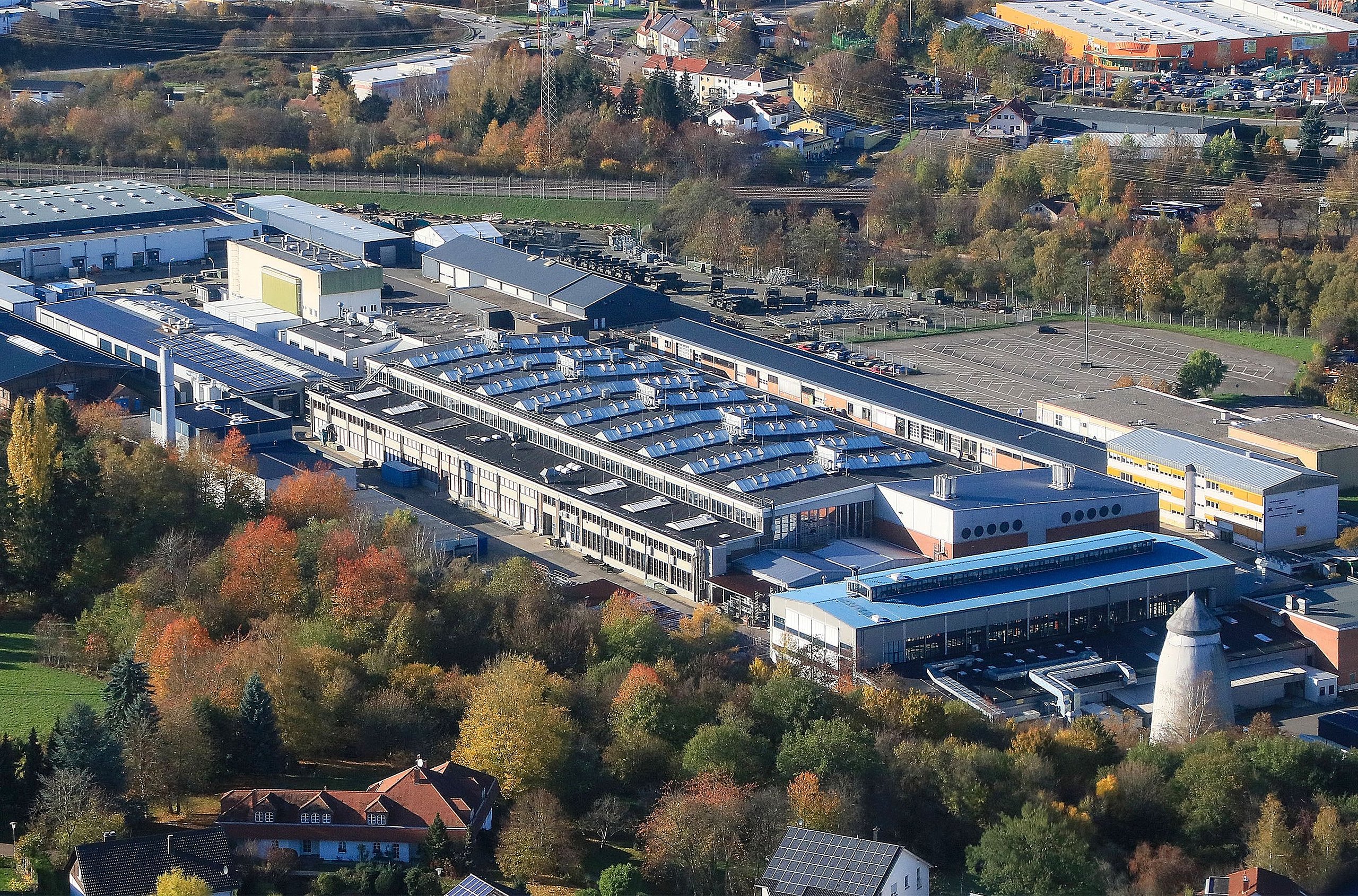 Luftbild des St. Wendeler Werks der Heeresinstandsetzungslogistik. In den Dächern einiger Gebäude spiegelt sich die Sonne. Die Anlage ist umgeben von bunt belaubten Bäumen, dem Flusslauf der Blies und einer Gleisstrecke der Deutschen Bahn.
