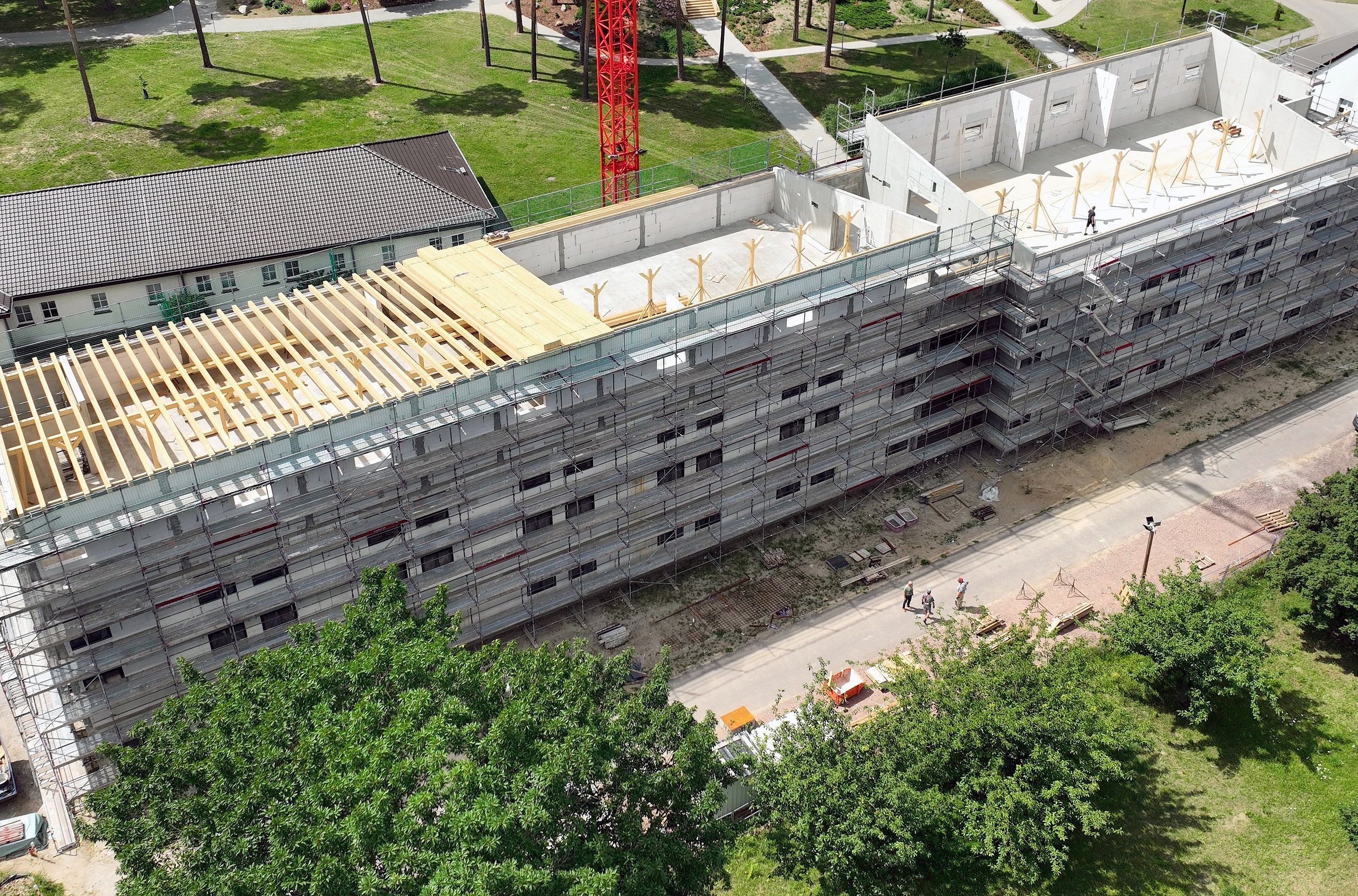 Luftaufnahme eines mehrstöckigen Gebäudes im Rohbau mit eingerüsteter Fassade und teilweise installiertem Dachstuhl in einer begrünten Umgebung.